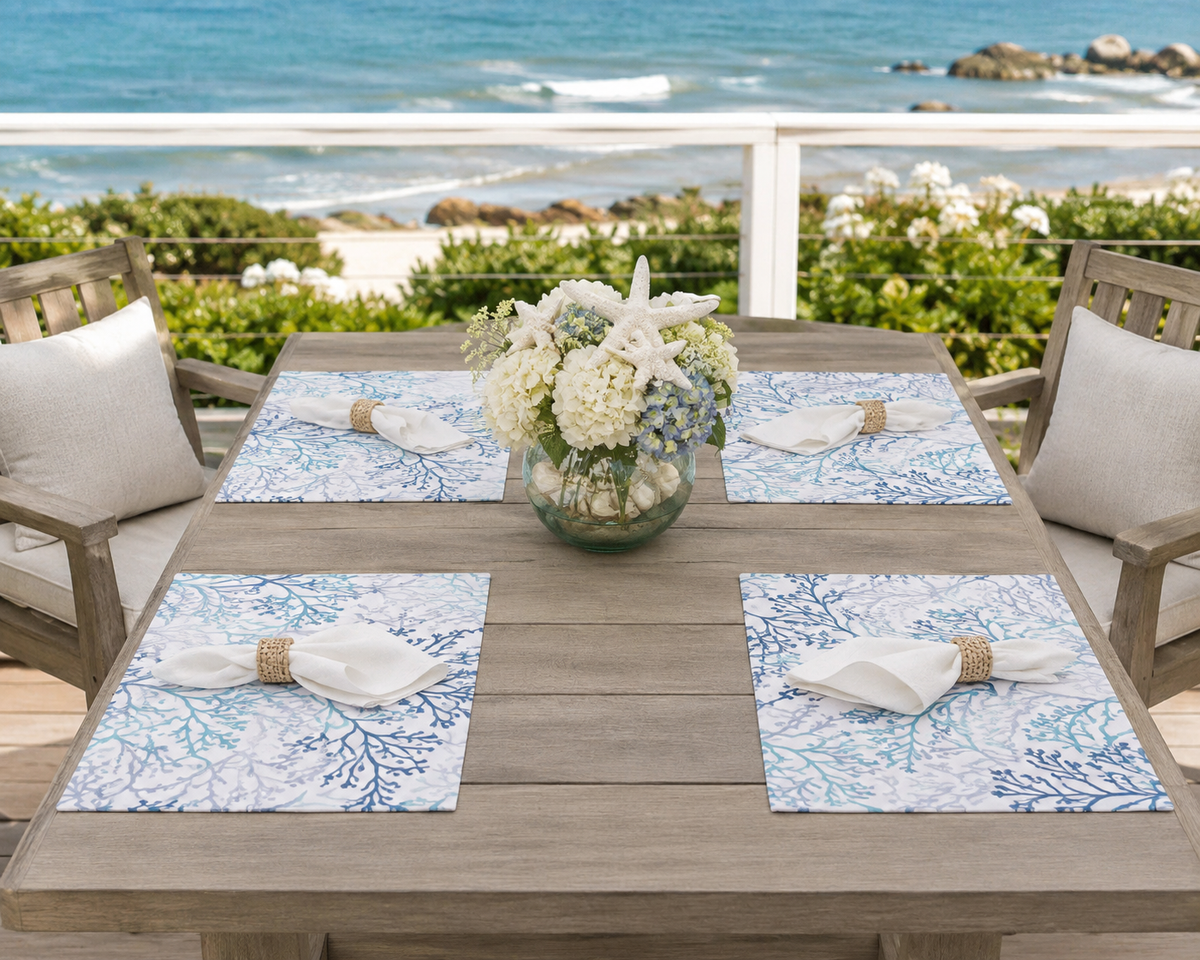 Four rectangular outdoor table placemats by Rennie and Rose with a blue coral branch pattern on a white background are arranged around a wooden dining table set for four, with a floral centerpiece and beach chairs, overlooking an ocean view.